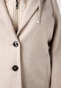 Beige wollen blendjas met een gestructureerd ontwerp, voorzien van een rits, een gewatteerde binnenvoering en drie zwarte knopen voor sluiting.