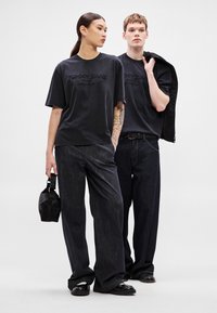 Tricouri grafice negre cu logo-ul "TOMMY JEANS", asortate cu pantaloni din denim închis la culoare. Femeia poartă o geantă mică neagră; amândoi poartă încălțăminte închisă la culoare.