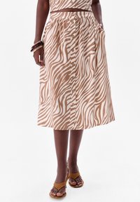 Jupe mi-longue en imprimé zèbre beige et marron, dotée d'un devant boutonné, d'une taille élastique et de poches latérales. Associée à des sandales beiges.