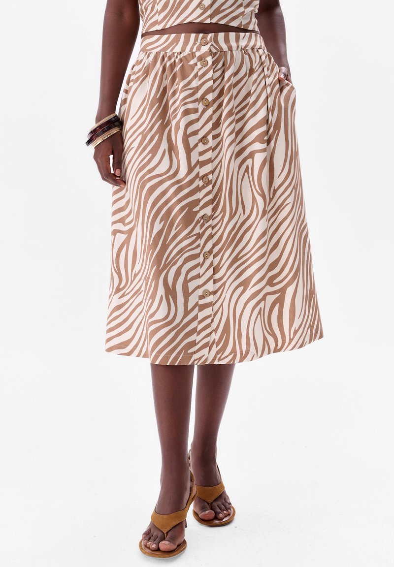 Jupe mi-longue en imprimé zèbre beige et marron, dotée d'un devant boutonné, d'une taille élastique et de poches latérales. Associée à des sandales beiges.