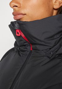 Veste isolante noire avec un col haut et une fermeture éclair rouge contrastante. Le tissu a une apparence lisse et texturée avec des coutures.