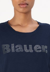 Marineblå t-skjorte med rund hals, prydet med sølvfargede rhinestone-bokstaver som staver "Blauer" i en leken skrifttype.