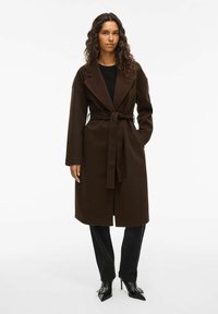 Manteau marron ceinturé avec un col cranté, des manches longues et une texture lisse, associé à un pantalon noir et des escarpins à bout pointu.