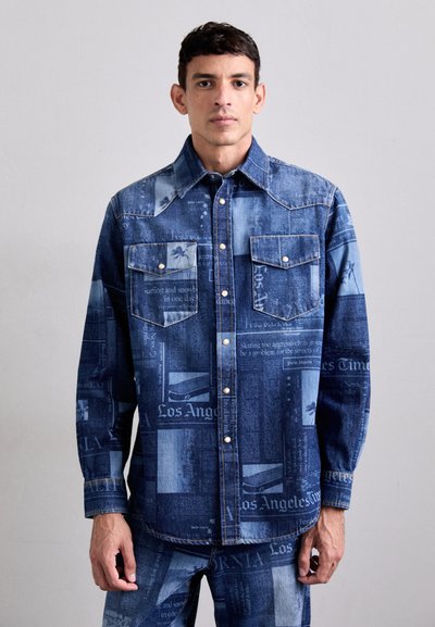 La chemise en denim présente une palette de couleurs bleues avec des motifs d'imprimés de journaux. Elle comprend des poches poitrine et une fermeture à boutons-pression, offrant un design texturé.