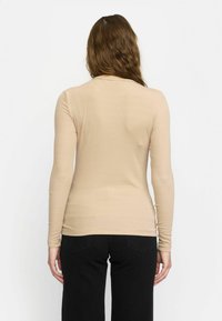 Beige hosszú ujjú ing, puha, rugalmas anyagból készült. Kerek nyakkivágással és testhez simuló fazonnal rendelkezik, fekete nadrággal párosítva.