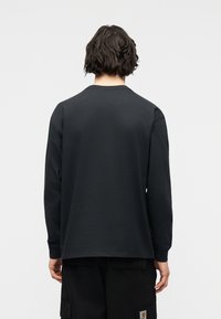 Άτομο με κοντά κυματιστά μαλλιά, ντυμένο με απλό μαύρο πουκάμισο μακρυμάνικο και μαύρο παντελόνι, στέκεται με την πλάτη προς τα εμπρός σε ουδέτερο φόντο.