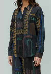 Persona che indossa una camicia a maniche lunghe con bottoni e pantaloni con motivi geometrici astratti multicolori in tonalità scure.