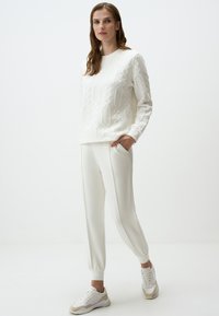 Witte getextureerde trui met een geometrisch patroon, gecombineerd met witte tapered broek, afgewerkt met witte en beige sneakers.