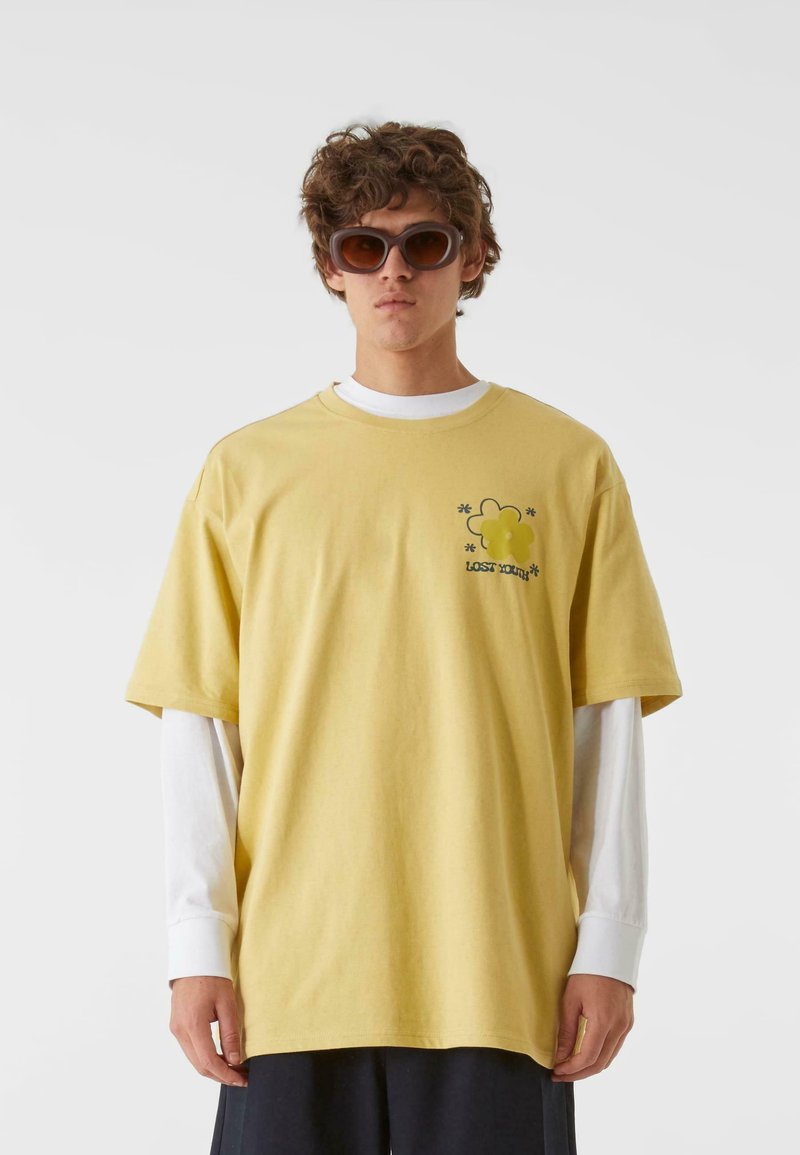Ung person iført en overdimensioneret gul T-shirt over hvide lange ærmer og store brune solbriller, stående mod en ensfarvet baggrund.