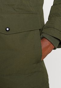 Olivgrön parkajacka med en knäppt bröstficka, ribbade muddar och synliga sömnadsdetaljer som visar en slitstark tygkvalitet.