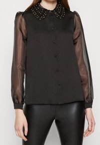 Svart blus med en dekorativ pärlkant, med genomskinliga långa ärmar och en knappframat. Tyget är Slätt med en lätt lyster.