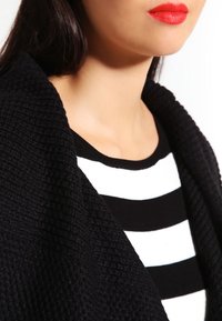 Svart texturerad cardigan över en svart och vit randig skjorta. Läppstiftet är knallrött och framhäver halsringningen.