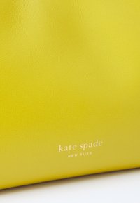 Žuta kožna torba s glatkom teksturom koja s prednje strane sadrži reljefni logo "kate spade NEW YORK" u bijeloj boji.