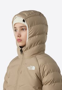 Noor inimene kannab beeži värvi puffer-jopet kapuuts peas ja valget kootud mütsi, näoga vasakule, neutraalne ilme.