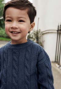 Donkerblauwe kabelgebreide trui gemaakt van een zachte, gestructureerde stof. Voorzien van een ronde hals en geribbelde manchetten, gedragen door een lachend kind.