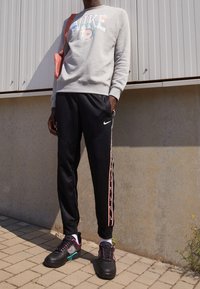 Person som bär en grå Nike-tröja, svarta Nike träningsbyxor med sidostrippar, svarta sneakers med rosa och turkosa detaljer, stående utomhus.