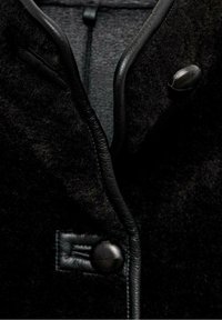 Gros plan sur un manteau en laine noire avec garniture en cuir montrant deux boutons recouverts de cuir noir et un tissu texturé.