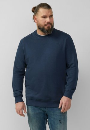 Donkerblauwe sweatshirt met een ronde hals, lange mouwen en ribgebreide manchetten. Gecombineerd met vervaagde blauwe jeans, die een ontspannen pasvorm tonen.