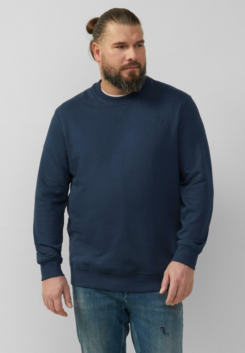 Le sweat-shirt bleu marine présente un col rond, des manches longues et des poignets côtelés. Associé à un jean bleu délavé, offrant une coupe décontractée.