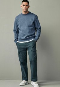Sweatshirt albastru cu guler rotund, pantaloni cargo verzi cu buzunare laterale și adidași albi. Ținută casual cu o croială lejeră și un design minimalist.