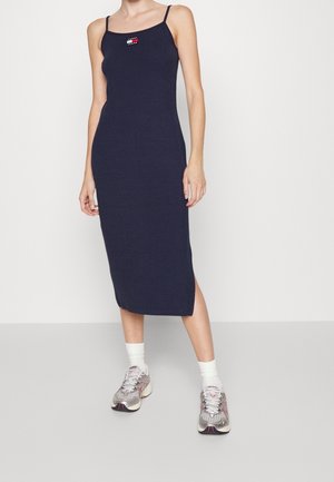 Kobieta ubrana w granatową sukienkę midi na ramiączkach spaghetti, z logo Tommy Jeans na piersi, białe skarpetki i srebrno-szare trampki.
