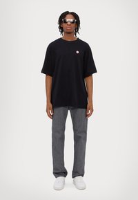 Black oversized T-shirt with a small red logo, paired with gray straight-leg jeans and white sneakers, worn with dark sunglasses.