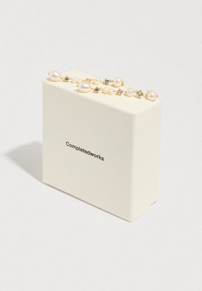 Joalharia em tons dourados com pérolas e cristais, exibida numa caixa de cor creme rotulada "Completedworks". Texturas suaves e design elegante.