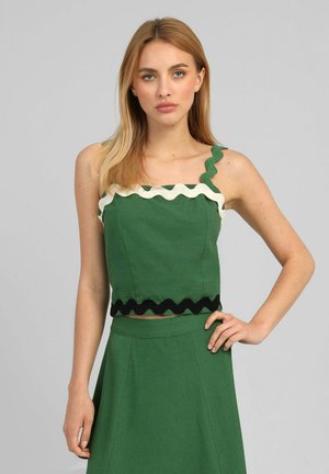 Femme debout, la main sur la hanche, portant un haut sans manches vert et une jupe assortie, toutes deux ornées d'un bord ondulé noir et blanc.