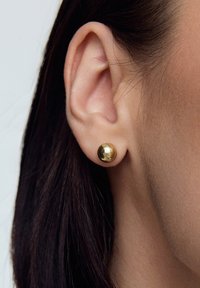 Złota kolczyk wkręcany o gładkiej, lustrzanej powierzchni, pokazany na uchu. Zbliżenie pokazuje okrągły kształt i błyszczącą fakturę.