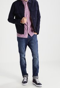 Giacca a zip blu sopra una camicia a quadri rossi e bianchi, jeans blu e sneakers nere con suole e lacci bianchi.