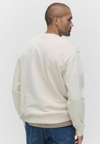 Jasnobeżowy sweter z okrągłym dekoltem, długimi rękawami i gładką teksturą. Posiada ściągacze na rękawach i u dołu, zestawiony z niebieskimi dżinsami.