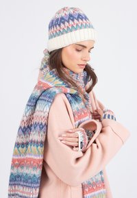Giovane donna che indossa un cappotto rosa pastello, un cappello a fantasia multicolore, una sciarpa coordinata e guanti senza dita, che guarda in basso con gli occhi chiusi.