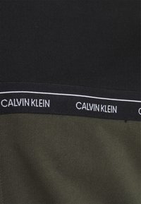 Ingrandimento di un tessuto nero e verde oliva con una vita nera caratterizzata da un marchio bianco "Calvin Klein" e una sottile striscia bianca.