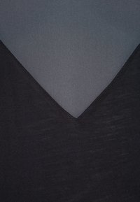 Černý top s výstřihem do V, který má průhledný šedý overlay v oblasti dekoltu, vystihující hladkou texturu a vrstvený design.