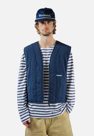 Mężczyzna noszący granatową czapkę z napisem „Weekend.”, granatową pikowaną kamizelkę na białej i granatowo-pasiatej koszuli z długim rękawem oraz beżowe spodnie.