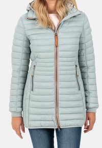 Mujer vistiendo una chaqueta acolchada con capucha de color azul claro, con cremallera frontal y bolsillos laterales con cremallera, combinada con jeans azules.