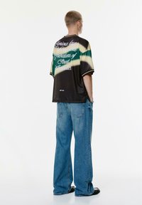 Zwart, groen en beige tie-dye shirt met korte mouwen met tekst op de achterkant, gecombineerd met lichtblauwe wijde jeans en zwarte schoenen.