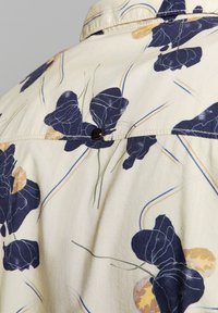 Lichtbeige shirt met marineblauw bloemenpatroon en gele accenten. Voorzien van een achterste knoop, gladde stof en kraagontwerp.