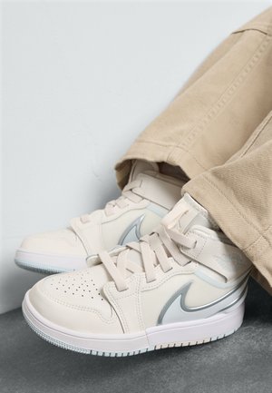 Lysbeige sneakers i syntetisk materiale, med perforeringer, sølvaccenter og hvide såler. Båret med beige bukser.