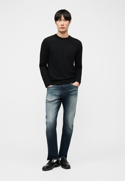 Jeune homme debout, les mains dans les poches, portant une chemise noire à manches longues, un jean bleu délavé et des chaussures en cuir noir sur un fond blanc.