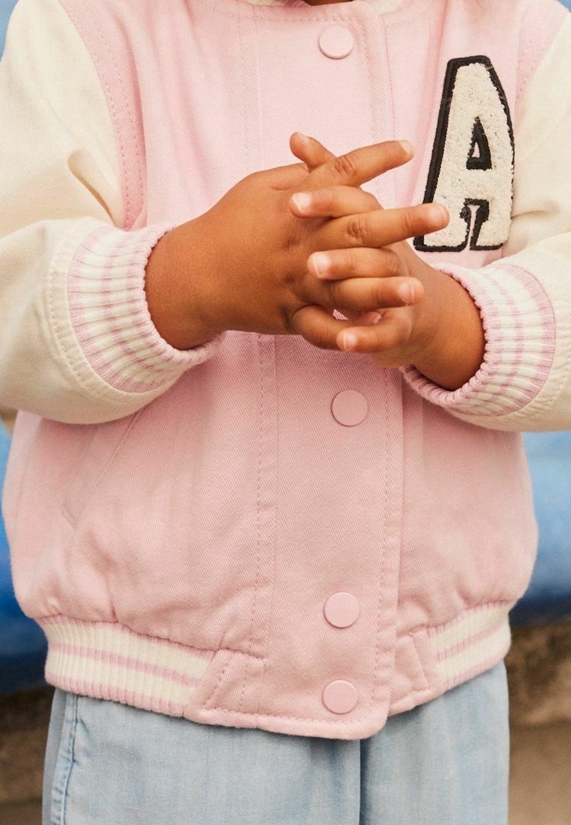 Enfant portant une veste rose et blanche avec un écusson lettre A, joignant les mains devant un pantalon bleu clair.