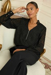 Bluză neagră cu un model texturat în valuri, guler cu fermoar și mâneci pliate, purtată cu pantaloni închisi la culoare. Scaunul este dintr-un material deschis la culoare.