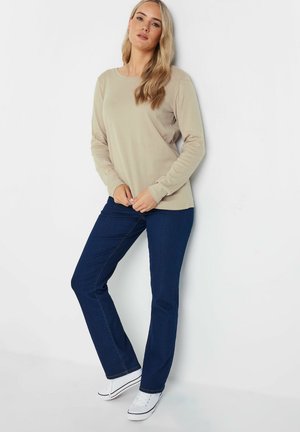 Beige langslijfshemd met een klassieke ronde hals, gecombineerd met donkerblauwe jeans. Model draagt witte sneakers, met de handen op de heupen geplaatst.