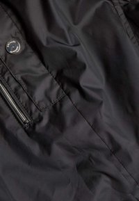 Zwarte nylon jas met een ritssluiting en een drukknoopdetail. Glad textuur met een minimalistisch ontwerp en geen zichtbare patronen.