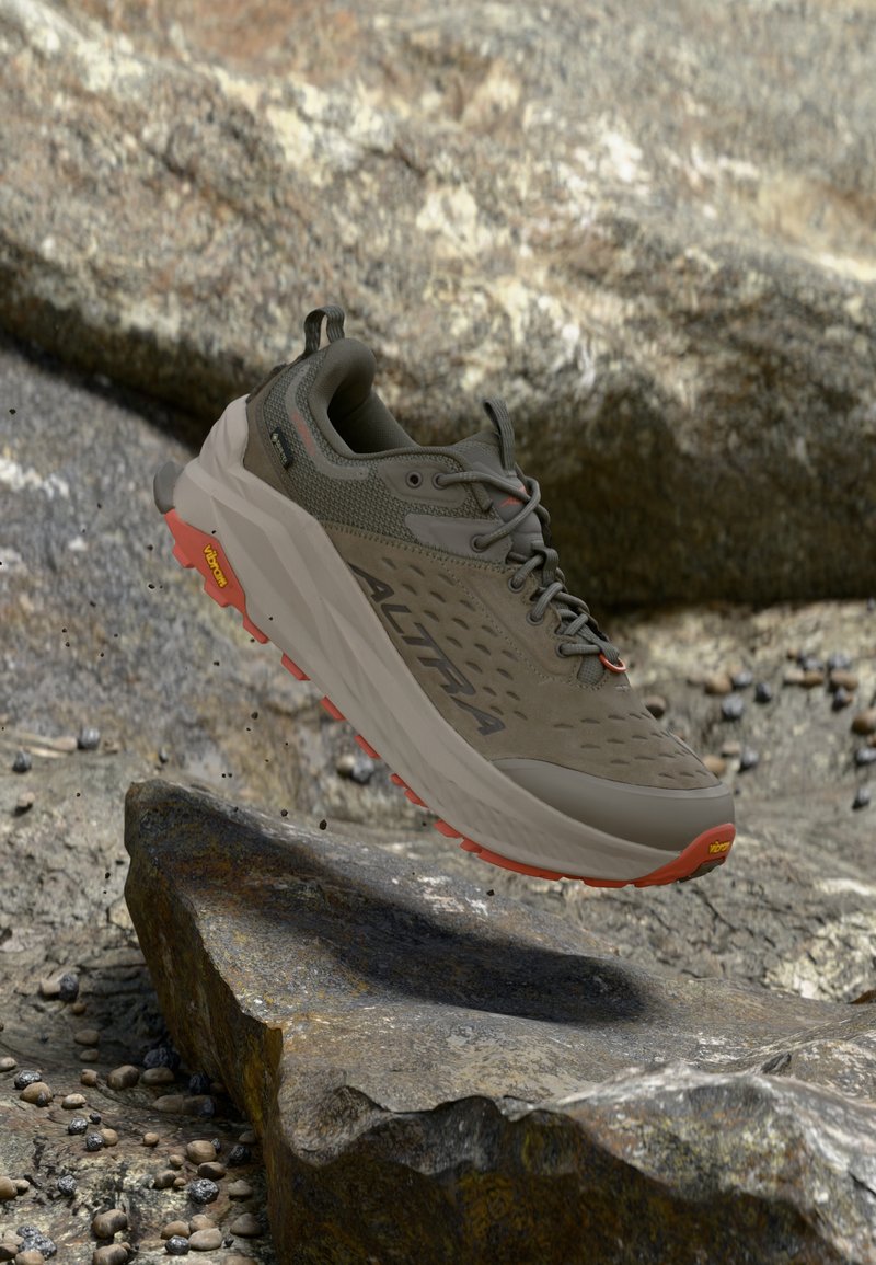 Καφέ και γκρι πεζοπορικό παπούτσι με κόκκινη σόλα Vibram να αιωρείται πάνω από πετρώδες έδαφος.