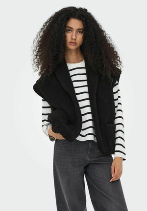 Sort, fuzzy vest med bred krave og uden ærmer, båret over en hvid stribet langærmet skjorte. Løse grå jeans og hænderne foldet.