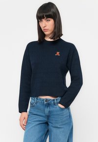 Granatowy sweter z dzianiny o prążkowanej teksturze i skróconym kroju. Posiada haftowane logo misia po lewej stronie klatki piersiowej. Długie rękawy.