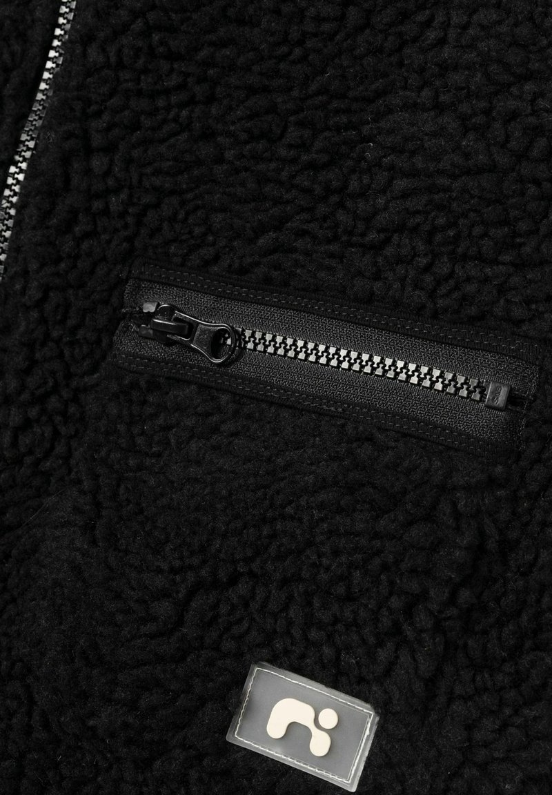Tissu polaire noir texturé avec une poche zippée noire horizontale et un patch logo cousu en dessous.