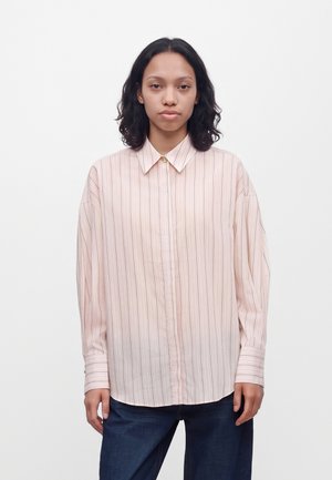 Femme aux cheveux foncés mi-longs portant une chemise ample rose pâle rayée à boutons et un jean bleu foncé, debout devant un fond uni.