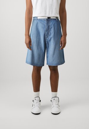 Personne portant un short en jean bleu clair, des chaussettes blanches, des baskets blanches et noires, et un haut blanc sans manches sur un fond gris uni.